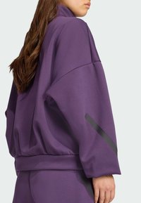Fialová športová mikina s vysokým golierom, spadnutými ramenami, textúrovanou látkou a čiernymi akcentmi na rukávoch.