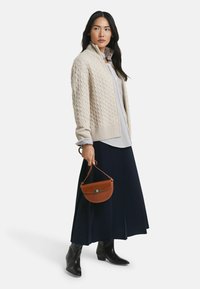 Femme portant un pull zippé en tricot torsadé crème, une blouse légère, une jupe midi bleu marine, des bottes noires, tenant un sac à main demi-lune marron par sa anse.