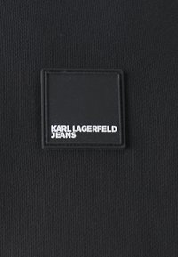 Tessuto nero con una finitura texture, presenta una toppa quadrata nera con la scritta "KARL LAGERFELD JEANS" in caratteri bianchi.
