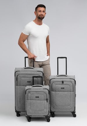 Trois valises en tissu gris de tailles variées avec des poignées et des roues noires. Chacune possède une poche frontale et des accents cousus.