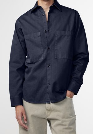 Camisa de manga larga azul marino hecha de un tejido suave, con cuello, dos bolsillos frontales y botones negros a lo largo del frente.