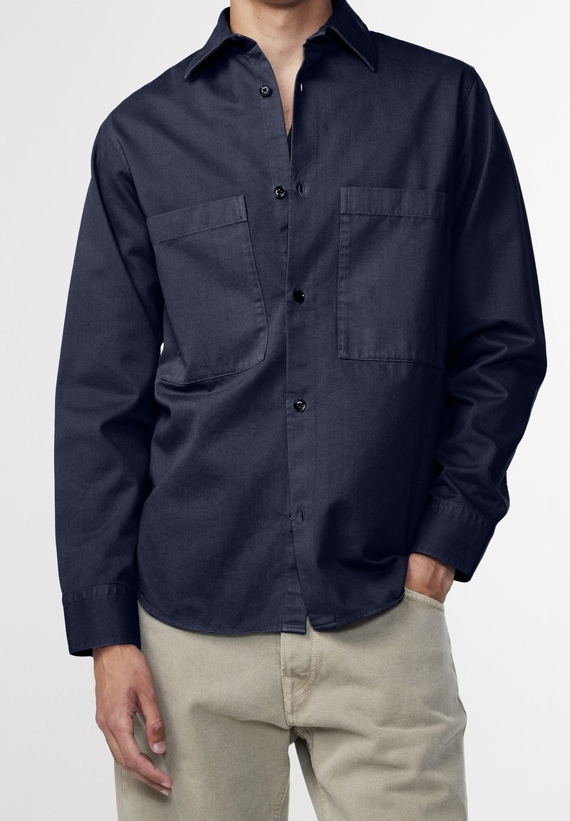 Camicia a maniche lunghe blu navy realizzata in tessuto liscio, dotata di colletto, due tasche frontali e bottoni neri lungo la parte anteriore.