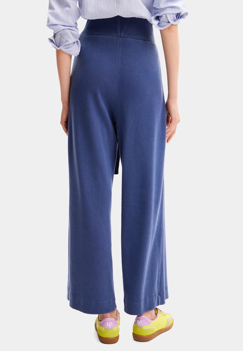 Persona che indossa pantaloni azzurri a vita alta e larghi, una camicia azzurro chiaro arrotolata e sneakers gialle con dettagli rosa, in piedi contro uno sfondo bianco.