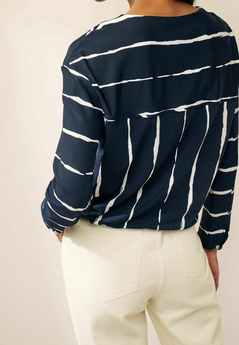 Femme portant un haut à manches longues bleu marine avec des rayures blanches irrégulières, associé à un pantalon taille haute blanc cassé, vue de dos.