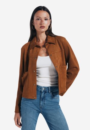 Giacca in suede marrone con colletto, bottoni a pressione e due tasche frontali. Indossata sopra una canotta bianca e jeans blu. Texture liscia.