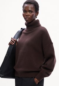 Maglione a collo alto marrone lavorato a maglia con una vestibilità morbida, polsini e orlo a coste. Indossato con una borsa a spalla in pelle nera, caratterizzata da una texture liscia.