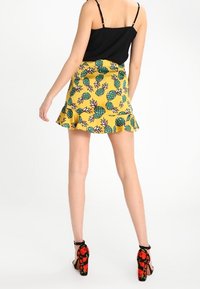 Jupe jaune avec un imprimé ananas, ornée d'un ourlet volanté ; assortie à un top à bretelles noir et à des chaussures à talons noirs avec des accents floraux.