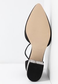 Sandale noire à bout pointu avec des lanières fines, une semelle beige texturée, et un talon bloc orné de petits strass argentés.