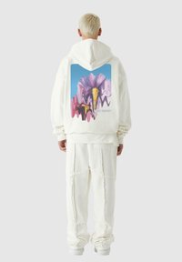 Biely mikina s farebnou kvetinovou grafikou na chrbte, ktorá obsahuje odtiene ružovej a fialovej, v kombinácii s ladiacimi bielymi nohavicami.