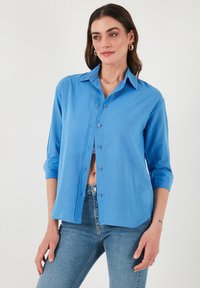 Camicia button-up azzurro chiaro con colletto e maniche arrotolate, realizzata in un tessuto liscio e strutturato, con chiusura a bottoni classica.
