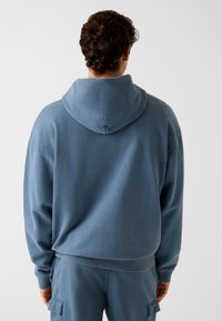 Bluza z kapturem w miękkim niebieskim materiale. Luźny krój z prążkowanymi mankietami i dołem. Posiada kieszeń kangura oraz bezszwowy tył.