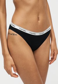 Γυναίκα με μαύρο εσώρουχο τύπου μπικίνι Calvin Klein με λευκή branded ζώνη, δείχνοντας τη μέση, το τατουάζ στο ισχίο και τα χέρια στα πλευρά.