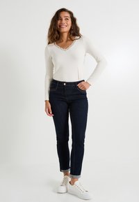 Femme souriante, portant un haut à manches longues blanc avec un col en V en dentelle, un jean bleu foncé retroussé et des sneakers plateformes blanches, se tenant devant un fond blanc.