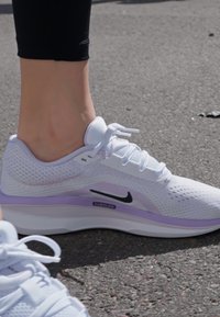 Hvit atletisk sko med mesh-overdel, lilla aksenter og svart Nike-logo. Har snørebånd og polstret såle for komfort.
