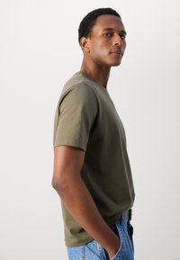 Olivenfarvet bomulds T-shirt med rund hals, korte ærmer og en afslappet pasform. Båret med blå denim jeans med synlige stikninger.