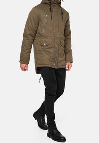 Veste isolante vert olive avec fermeture éclair à l'avant, double poches poitrine et capuche ajustable avec cordon de serrage. Associée à un pantalon noir et des bottes.