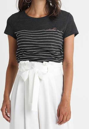 Vrouw in een zwart-wit gestreepte t-shirt met geborduurd "love" en hoge witte broek vastgemaakt met een grote strikriem.