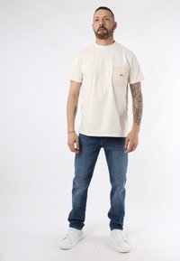 T-shirt in cotone beige con tasca a righe sul lato sinistro, abbinata a jeans blu e scarpe da ginnastica bianche, in piedi su uno sfondo neutro.