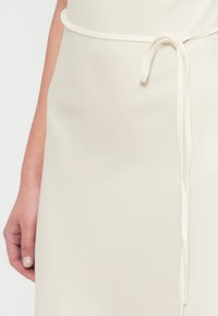 Λευκό, ρέον φόρεμα με ομαλή, υφασμάτινη υφή και λεπτομέρεια δέσιμου στο πλάι στη μέση, εμφανίζεται εν μέρει με ένα χέρι που το κρατάει.