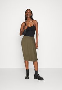 Čierny top bez rukávov s zberom, olivovo zelená midi sukňa a čierne členkové topánky. Modelka drží čiernu tašku; má elegantný, minimalistický dizajn.