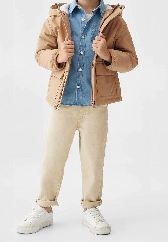 Giacca con cappuccio tan, con tasche frontali, camicia azzurro chiaro con bottoni, pantaloni beige e sneakers bianche. Tessuti morbidi e vestibilità casual.