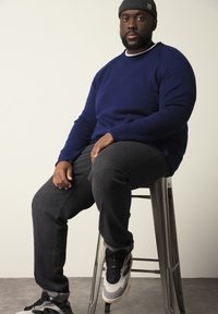Pull en tricot bleu marine, jean gris foncé et chaussures de sport noires/grises portés par une personne assise sur un tabouret en métal.