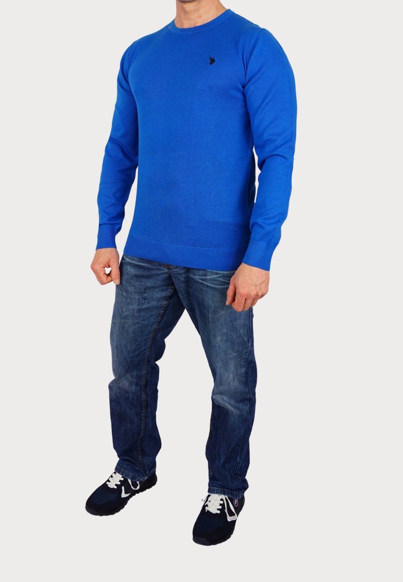 Homme portant un pull bleu vif à manches longues, un jean bleu et des baskets noires avec des lacets blancs, debout devant un fond uni.