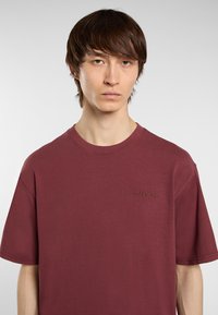 Mladý muž se rovnými hnědými vlasy, oblečený v jednoduchém vínovém tričku s malou vyšívanou nášivkou na hrudi, na bílém pozadí.