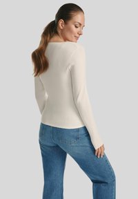 Kobieta z długimi brązowymi włosami związanymi w kucyk, ubrana w dopasowaną białą bluzkę z długim rękawem i niebieskie spodnie z wysokim stanem, stojąca na tle jednolitego tła.