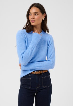 Kvinde, der kigger sidelæns iført en lyseblå sweater, mørke jeans med brunt bælte og sølvøreringe mod en ensfarvet hvid baggrund.