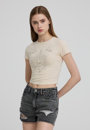 T-shirt cropped beige con maniche corte, decorata con una grafica grigia di rose e uccelli. Abbinata a pantaloni shorts in denim nero strappati.