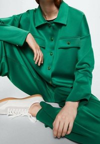 Zestaw zielonej satynowej koszuli i spodni z guzikami i kieszeniami, w połączeniu z białymi sneakersami z beżowymi akcentami. Gładka tekstura, nowoczesny design.