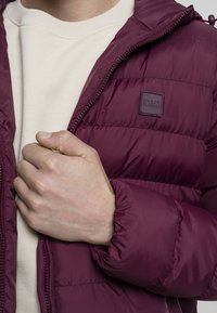 Bordowa kurtka puchowa z zamkiem z przodu, regulowanym kapturem i małą naszywką z logo. Posiada pikowany wzór oraz gładką, błyszczącą teksturę.