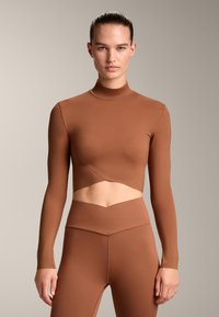 Bruine langemouw cropped top met hoge nek, met uitsnijding rond de taille. Glad, rekbaar materiaal met een aansluitende pasvorm.