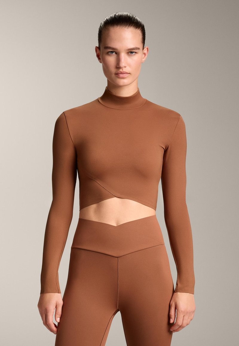 Bruine langemouw cropped top met hoge nek, met uitsnijding rond de taille. Glad, rekbaar materiaal met een aansluitende pasvorm.