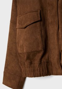 Détail d'une veste en daim marron avec une finition texturée, un ourlet élastiqué et une poche plaquée unique à l'avant.