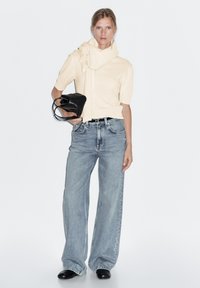 Maglione corto a maniche corte color beige chiaro con dettaglio sciarpa, abbinato a jeans a gamba larga blu chiaro a vita alta e stivali neri alla caviglia. Borsa nera.