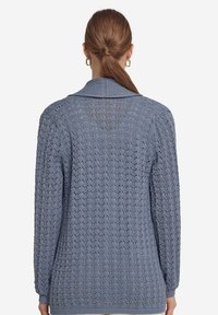 Ljust blått stickat kardiigan har ett strukturerat mönster, långa ärmar och en sjalkrage. Stickningen är något öppen och andas.