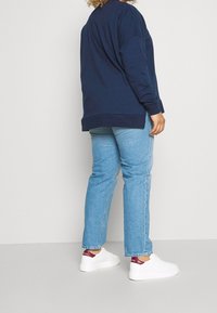 Felpa navy con maniche lunghe, vestibilità rilassata e spacchi laterali, abbinata a jeans azzurri chiari e sneakers bianche con dettagli bordeaux.