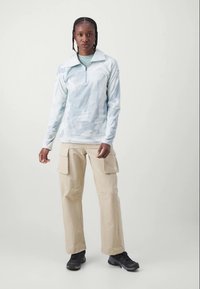 Donna con capelli intrecciati, che indossa un pullover azzurro chiaro a motivo, pantaloni cargo beige e scarpe nere, in piedi di fronte a uno sfondo leggero e uniforme.