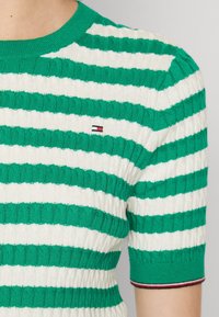 Maglione lavorato a maglia a righe verdi e bianche con maniche corte, texture a costine e un piccolo logo sul petto. Accenti rossi e blu ai polsini.