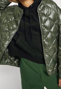 Prošívaná, lesklá, olivově zelená péřová bunda se zapínáním na patentky přes černou mikinu. Model má na sobě zelené kalhoty, čímž podtrhuje vrstvený vzhled.