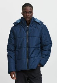 Mörkblå puffjacka med avtagbar huva, dragkedja framtill och elastiska ärmslut. Har horisontell sömnad och sidofickor.