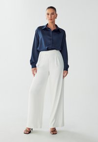 Blusa di satin blu navy con bottoni frontali e maniche lunghe, abbinata a pantaloni bianchi a gamba larga con vita alta e sandali neri con tacco.