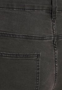 Black denim fabric with a smooth texture, featuring back pocket seams and stitching detail. Tailored fit suggested by shape.