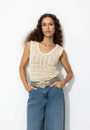 Débardeur en tricot beige avec un col en vagues, associé à un jean large en denim bleu taille haute et une ceinture en or tissée.
