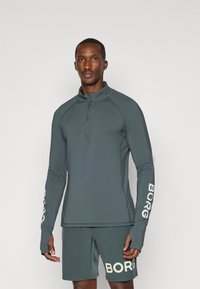 Αθλητική μπλούζα με μακριά μανίκια σε σκούρο τιρκουάζ, με φερμουάρ μέχρι το ένα τέταρτο, διαθέτει οπές για τους αντίχειρες και άσπρο λογότυπο "BORG" στα μανίκια. Συνδυάζεται με ταιριαστά σορτς.