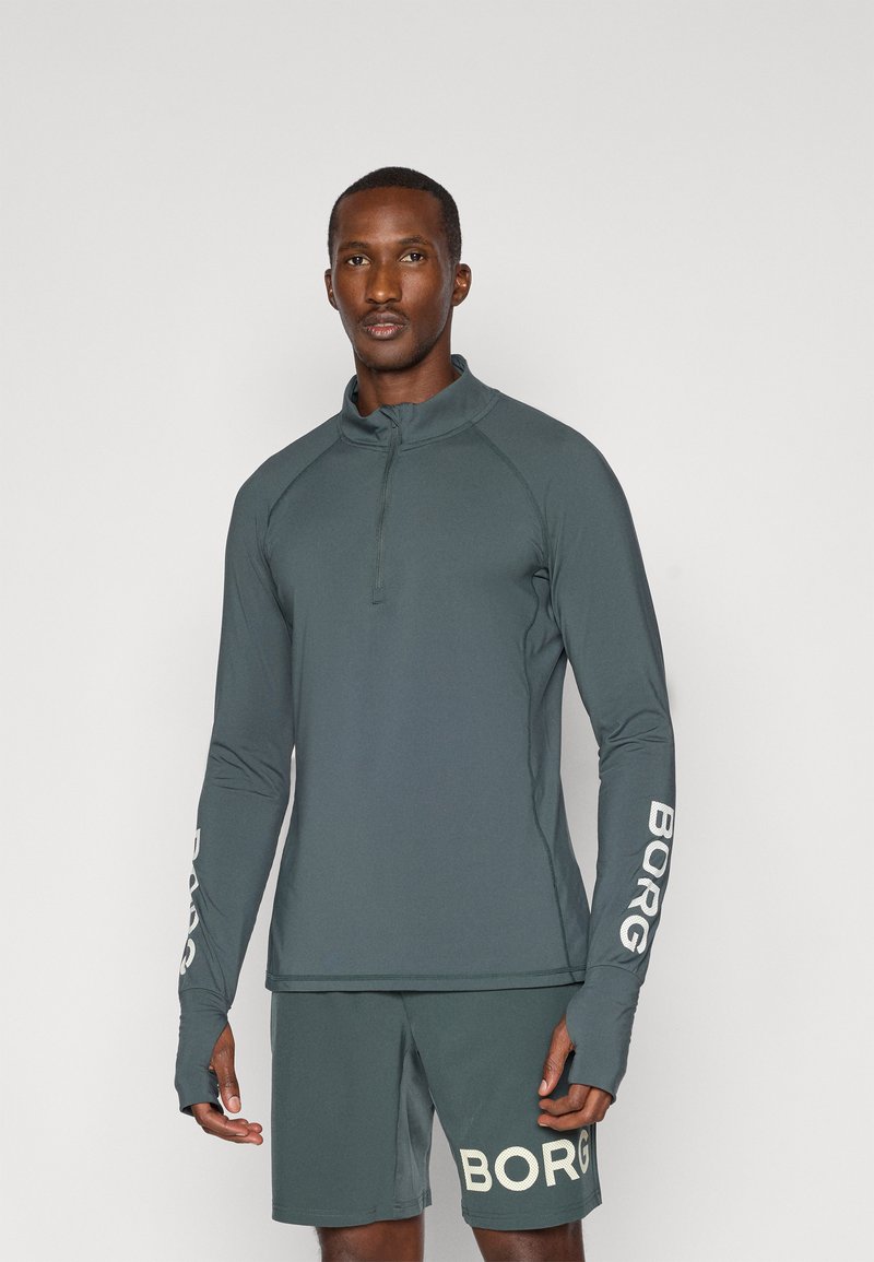 Αθλητική μπλούζα με μακριά μανίκια σε σκούρο τιρκουάζ, με φερμουάρ μέχρι το ένα τέταρτο, διαθέτει οπές για τους αντίχειρες και άσπρο λογότυπο "BORG" στα μανίκια. Συνδυάζεται με ταιριαστά σορτς.