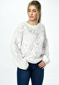 Biały sweter z dzianiny, zdobiony geometrycznym wzorem ażurowym, z szerokimi rękawami i ściągaczami, w połączeniu z ciemnoniebieskimi dżinsami.