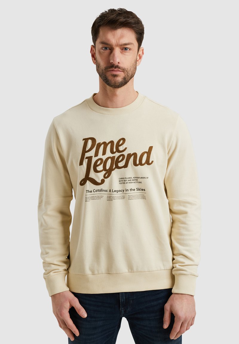 Sudadera color crema con cuello redondo. Presenta texto marrón en negrita y un estampado gráfico. Material suave, puños y dobladillo acanalados.